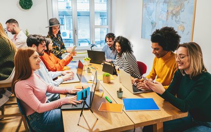 Junge stylische Leute sitzen gemeinsam im lichtdurchfluteten Raum am großen Tisch und nutzen verschiedene Devices wie Notebooks und Handys