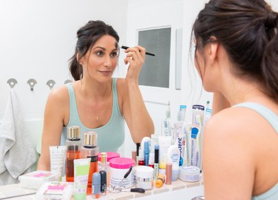 Junge Frau schminkt sich vor Badezimmerspiegel, vor dem Spiegel stehen diverse Kosmetikprodukte